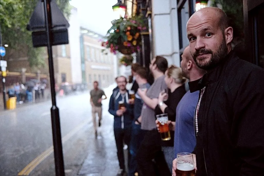 Ham looking into the camera, standing in front of a pub in London, rain pouring down in the background